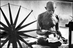 Gandhi at home next to a spinning wheel, which looms in the foreground as a symbol of India's struggle for independence